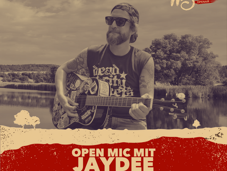 Jaydee steht mit seiner Gitarre am Obernzenner See und blickt fröhlich in die Menge seines Publikums. Er trägt eine Sonnenbrille.